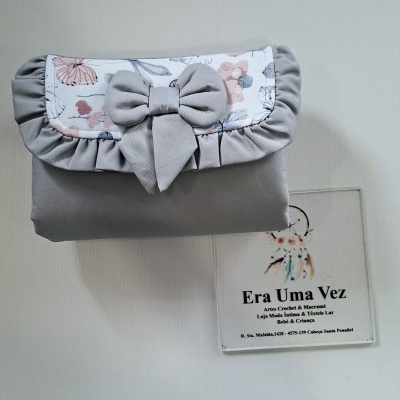 Bolsa cinza com aba estampada e laço, rótulo com texto ao lado.