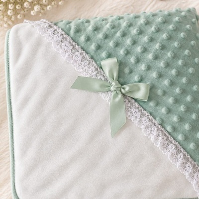 Manta de bebé dupla face branco e verde com renda e laço sobre superfície peluda com flores, ursinho e coração de madeira.