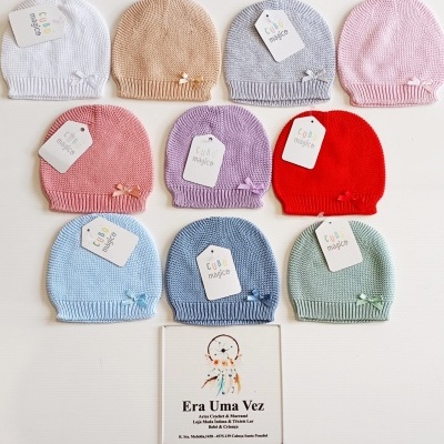 Dez gorros de malha coloridos para bebé com etiquetas Cuti Mojica, dispostos em fileiras e placa informativa com desenho de apanhador de sonhos.