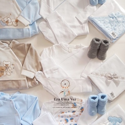 Roupas de bebé variadas em branco, azul claro e cinzento com bordados e mantas dobradas.