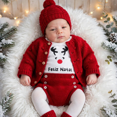 Bebé vestido com roupa de Natal vermelha e branca, sentado em superfície macia com decoração de Natal ao redor.