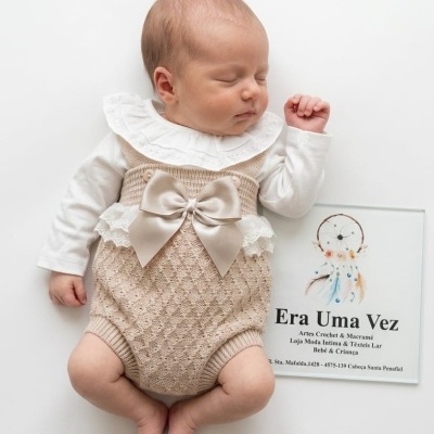 Bebé deitado com fato de malha bege com laço e body branco ao lado de cartaz Era Uma Vez