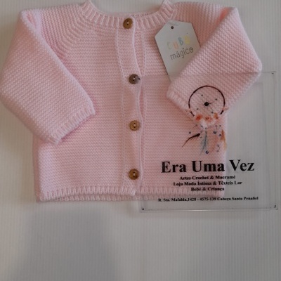 Casaco de bebé rosa em malha com botões de madeira, etiqueta pendurada e texto promocional em fundo branco