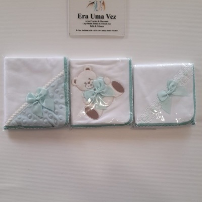 Três fraldas brancas para bebé com detalhes em azul claro e ursinho bordado, dispostas lado a lado sob uma placa 'Era Uma Vez'