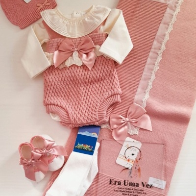 Conjunto de bebé cor-de-rosa com camisola e body branco, sapatinhos, meias, touca e manta com etiquetas visíveis.