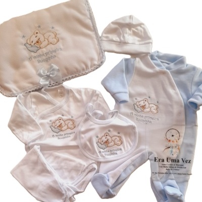 Conjunto de roupa de bebé com manta bege, body branco, babete e fato de dormir azul claro e branco com ursinho bordado e texto