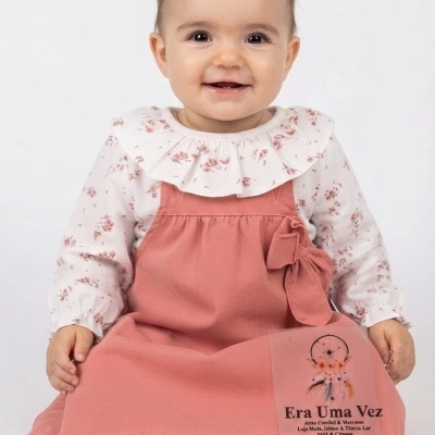 Bebé com vestido cor de rosa e camisa branca floral sentada em fundo branco.