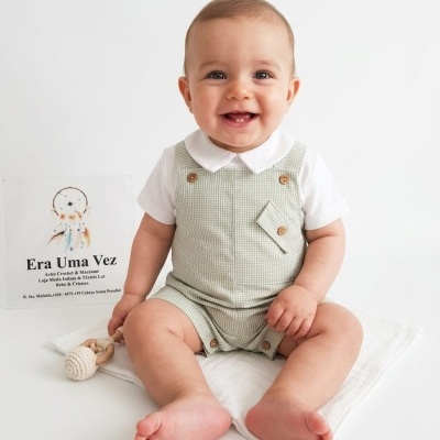 Bebé com jardineira verde claro, camisa branca e brinquedo de madeira sentado no chão