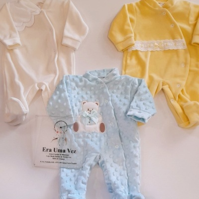 Pijamas de bebé branco, azul claro com ursinho e amarelo com faixa branca em fundo neutro.