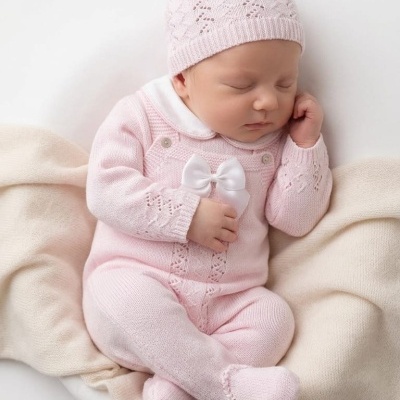Bebé vestido com fato e gorro de malha rosa deitado numa manta bege