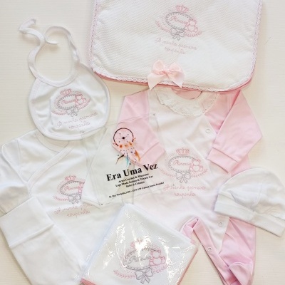 Conjunto roupa bebé branca e rosa com bordados e acessórios sobre fundo branco.