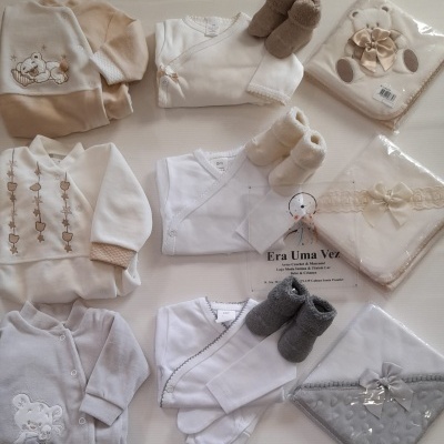 Conjunto de roupa e mantas de bebé em tons neutros com botins felpudos, dispostos sobre fundo branco com texto visível