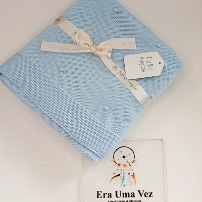 Manta azul clara tricotada com fita branca e etiqueta cubo mágico