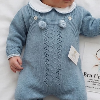 Bebé vestido com roupa azul de malha com padrão de renda e gorro, deitado sobre fundo branco.