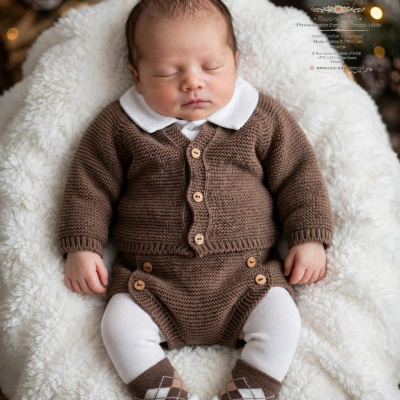Bebé a dormir vestido com camisola castanha de malha, collant branco, meias castanhas e sapatinhos brancos numa almofada branca.