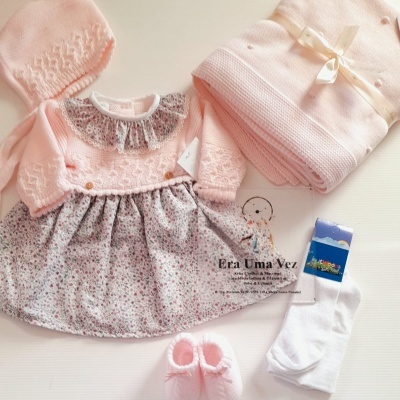 Conjunto de roupa para bebé menina com vestido floral, capuz e manta rosa, sapatinhos e meias brancas.