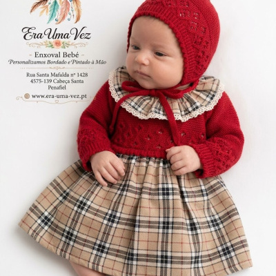 Bebé com vestido xadrez bege e corselet vermelho, gola de renda e gorro vermelho.