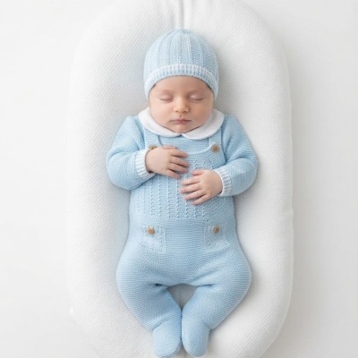 Bebé a dormir com macacão e gorro azul claro numa cama almofadada branca