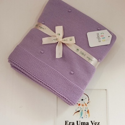 Manta lilás de malha com pompons e fita com etiqueta da marca Cubo mágico sobre fundo branco com placa informativa.