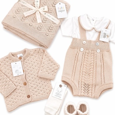 Conjunto de roupa de bebé com manta, casaco e ranita em malha bege rendada, body branco, meias brancas e sapatinhos em malha bege.