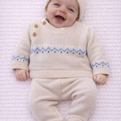 Bebé a sorrir vestido com conjunto bege de malha com padrão azul e botões de madeira deitado sobre superfície branca texturada