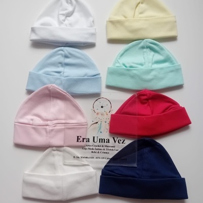 Oito gorros de bebé em algodão de várias cores dispostos em círculo com um cartão no centro sobre fundo branco