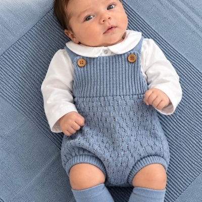 Bebé vestido com camisa branca e macacão azul de malha, meias azuis, deitado em manta azul.