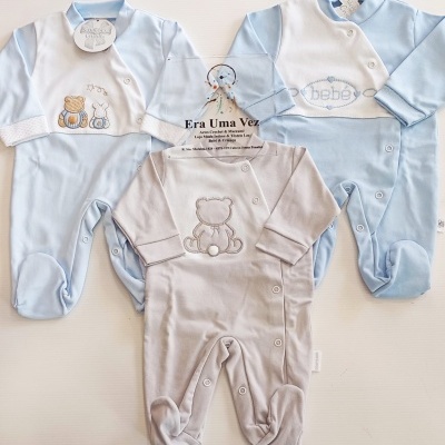 Três pijamas de bebé azul claro e bege com bordados e botões, etiquetas e cartão na mesa branca