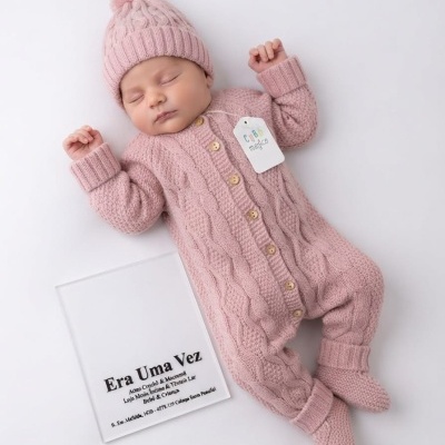 Bebé com fato de malha rosa trançada, gorro, botinhas e placa branca com texto