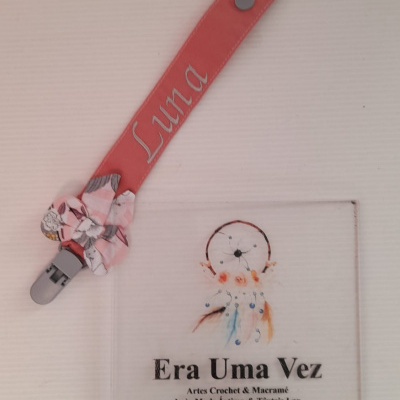 Presilha rosa com nome Luna e laço floral sobre mesa branca com cartão com texto e desenho.
