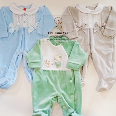 Três macacões de bebé em veludo azul, verde e bege com bordados e laços, sobre fundo branco e placa transparente com texto