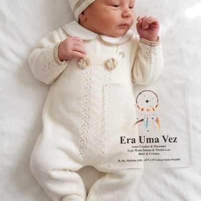 Bebé recém-nascido vestido com fato creme de tricô e gorro com detalhes castanhos, deitado numa superfície branca.