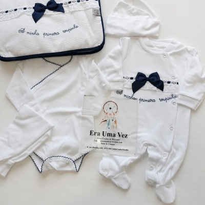 Conjunto de roupa de bebé branco com laço azul e manta com texto A minha primeira roupinha