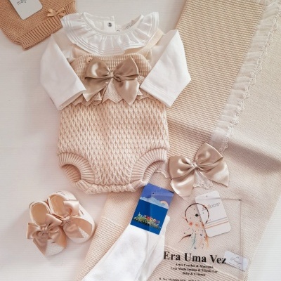 Conjunto de roupa de bebé bege e branco com body, meias, sapatinhos, gorro e manta detalhados com laços e etiquetas.