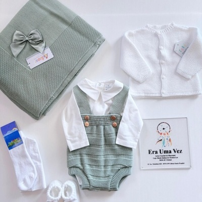 Conjunto de roupa para bebé composto por macacão verde, camisola branca, casaco branco, meias, sapatinhos e manta com laço verde.