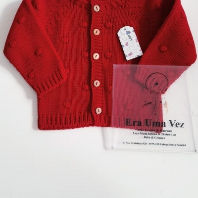 Casaco vermelho de malha com botões de madeira e saco plástico transparente com texto da loja