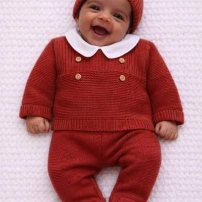 Bebé sorridente vestido com conjunto vermelho de malha e gola branca sobre manta branca.