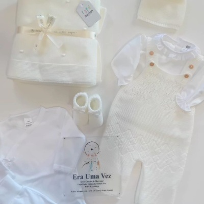 Conjunto de roupa e acessórios para bebé em branco sobre superfície branca, mostrando body, macacão, touca, sapatinhos, manta com ficha Cubo mágico e etiqueta Era Uma Vez