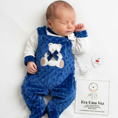Bebé vestido com macacão azul felpudo com ursinho bordado e chupeta branca, sobre fundo branco