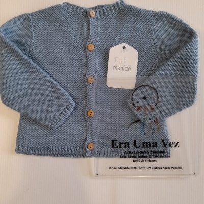 Casaco bebé azul com botões de madeira e etiqueta CUBO mágico sobre superfície clara com placa informativa.