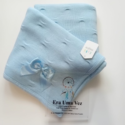 Manta de bebé azul clara com laço de cetim azul e etiqueta, sobre cartão transparente com texto da loja.