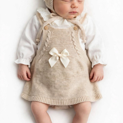 Bebé a dormir com gorro e vestido de malha bege com laços, e blusa branca.