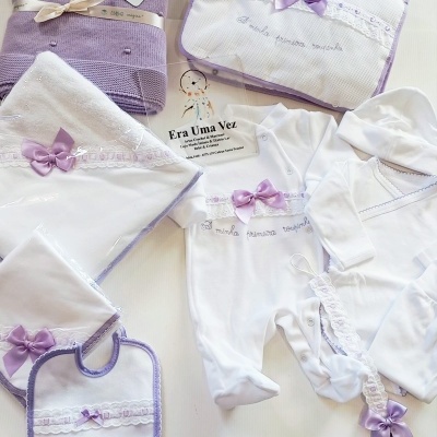 Conjunto de roupa e acessórios de bebé branco e lilás com laços de cetim e renda.