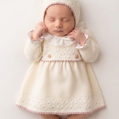 Bebé vestido com vestido branco em malha com renda e gorro branco com borda cor-de-rosa num fundo branco