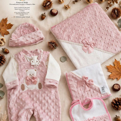 Conjunto de roupa e acessórios para bebé rosa com urso bordado e laços em tecido texturizado sobre pano bege com elementos naturais