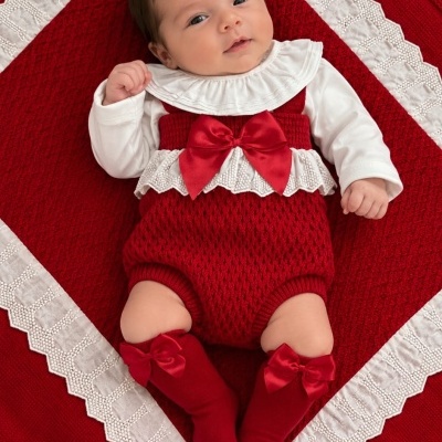 Bebé vestida com roupa vermelha e branca de tecido tricotado com laços, deitada em pano vermelho com borda branca rendada.