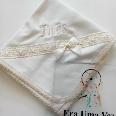 Manta de bebé branca com bordado e renda bege sobre papel com texto.