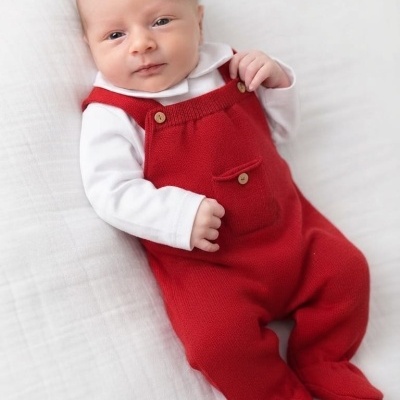 Bebé com macacão vermelho e camisola branca a deitar numa cama branca.