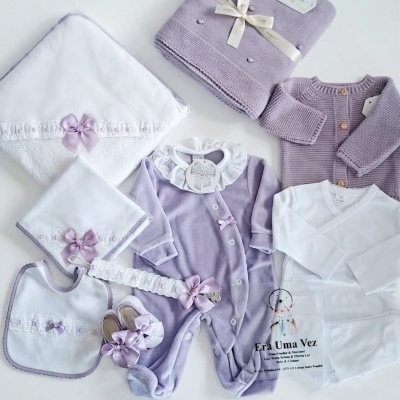 Conjunto bebé lilás e branco com pijama, casaco, manta, babete, sapatinhos e toalhas decorados com laços e rendas.