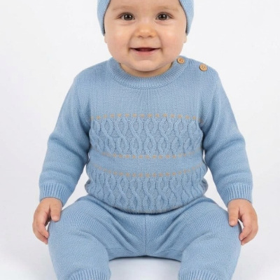 Bebé com roupa de malha azul e gorro com pompom sentado em fundo branco.